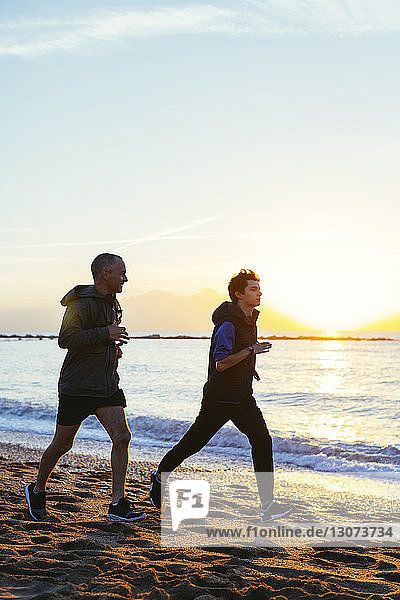 Confident father and son jogging on shore at beach against sky during sunset