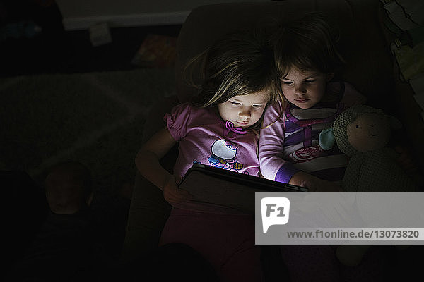 High angle view of sisters holding tablet computer while lying on sofa in darkroom at home