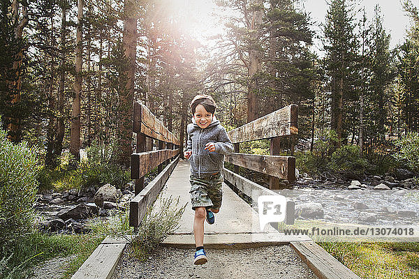 Portrait of boy running over wooden bridge against trees in forest
