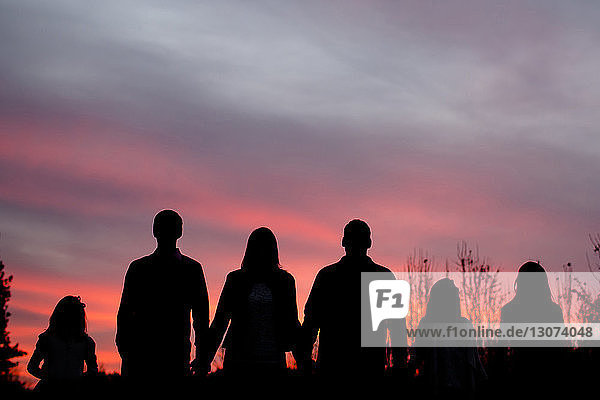 Scherenschnittfamilie hält sich an den Händen und steht bei Sonnenuntergang vor dramatischem Himmel auf dem Feld