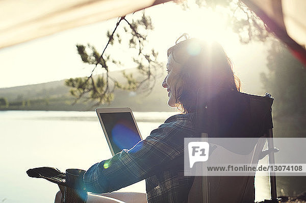 Smiling woman with tablet computer relaxing on armchair at lakeshore during sunny day
