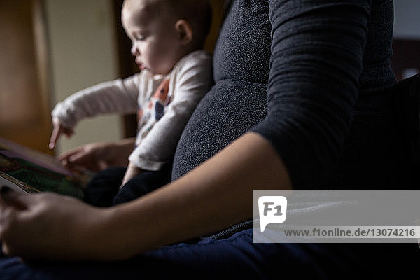 Mittelsektion einer schwangeren Frau  die zu Hause sitzend ihrer Tochter ein Bilderbuch zeigt