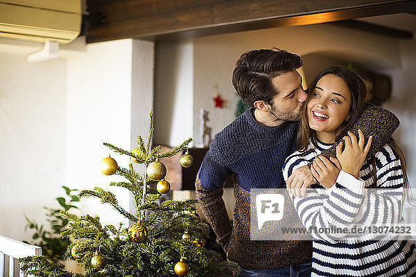 Mann küsst Freundin,  während er zu Hause am Weihnachtsbaum steht