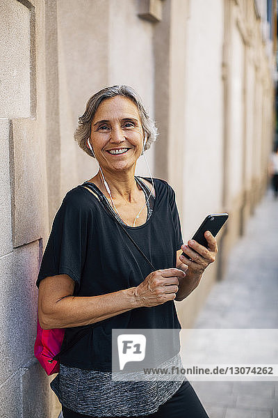 Porträt einer glücklichen Frau  die ein Smartphone in der Hand hält  während sie an der Wand steht