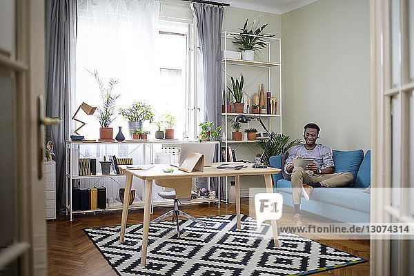 Glücklicher junger Mann benutzt Tablet-Computer  während er zu Hause auf dem Sofa sitzt