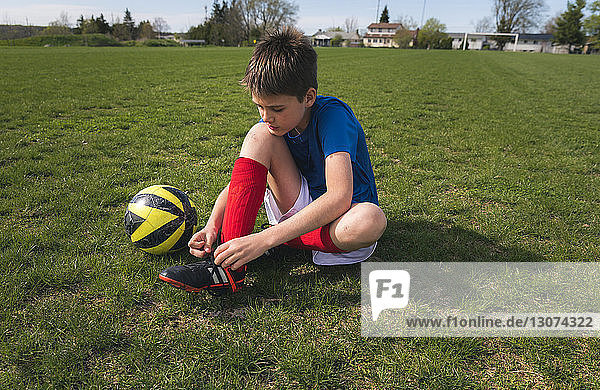 Junge in voller Länge beim Schnürsenkelbinden  während er neben einem Fussball auf einem Rasenplatz sitzt