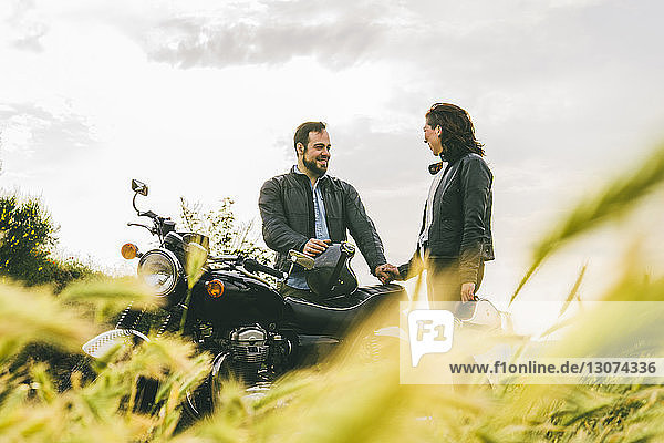 Couple talking while standing by motorcycle on grassy field against sky