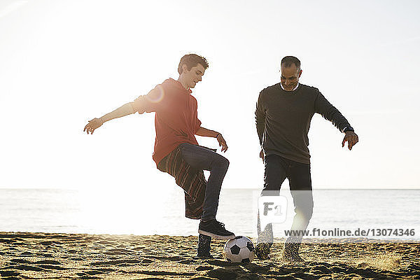 Playful father and son playing soccer at beach against clear sky during sunny day