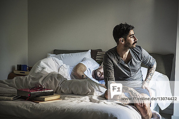 Thoughtful gay man looking away while partner sleeping on bed