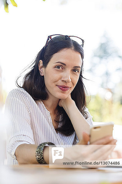 Portrait of confident woman holding smart phone while sitting at sidewalk cafe