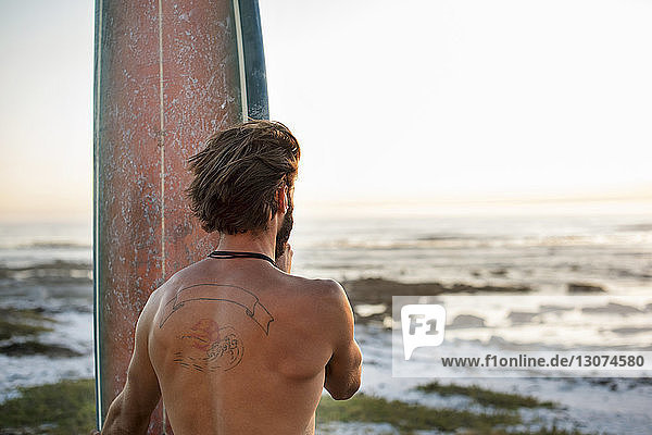 Rückansicht eines Mannes ohne Hemd mit Surfbrett am Strand bei Sonnenuntergang