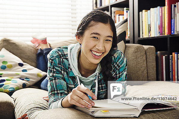 Porträt einer fröhlichen Teenagerin  die Hausaufgaben macht  während sie zu Hause auf der Couch liegt