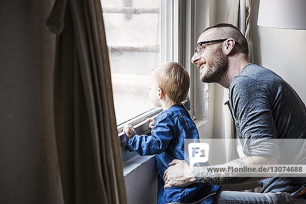 Glücklicher Vater mit Tochter  der durchs Fenster schaut  während er zu Hause sitzt