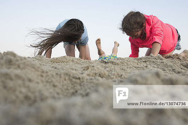 Ebenerdiges Bild von Schwestern  die mit Sand am Strand vor klarem Himmel spielen