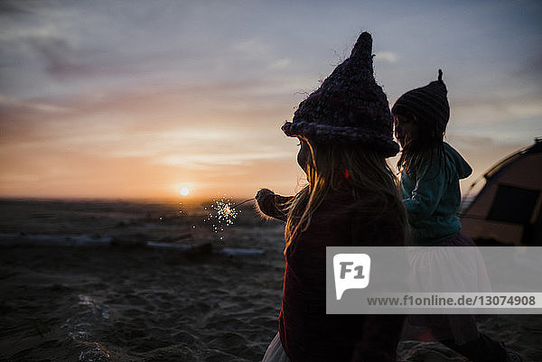 Schwestern spielen mit Wunderkerzen beim Camping am Strand während des Sonnenuntergangs
