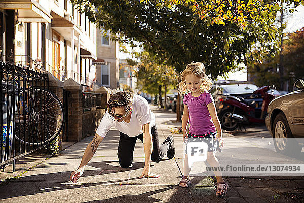 Girl looking down while father drawing hopscotch on footpath