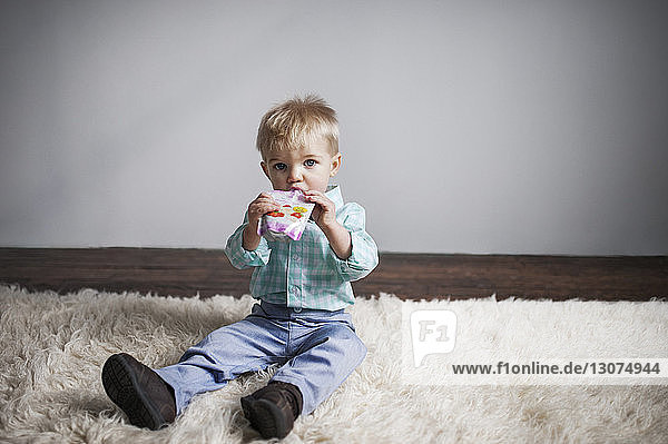 Porträt eines süßen Babyjungen  der zu Hause auf dem Teppich sitzend Saft aus der Packung trinkt