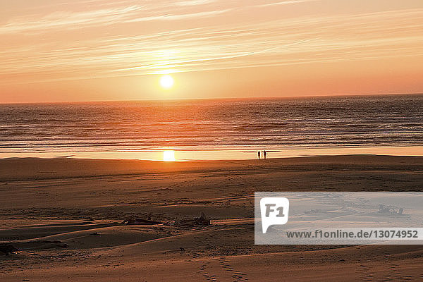 Entfernte Silhouette von Freundinnen  die bei Sonnenuntergang am Strand gegen den Himmel stehen