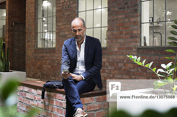 Geschäftsmann benutzt Smartphone  während er auf einem Sitz vor einem Bürogebäude sitzt