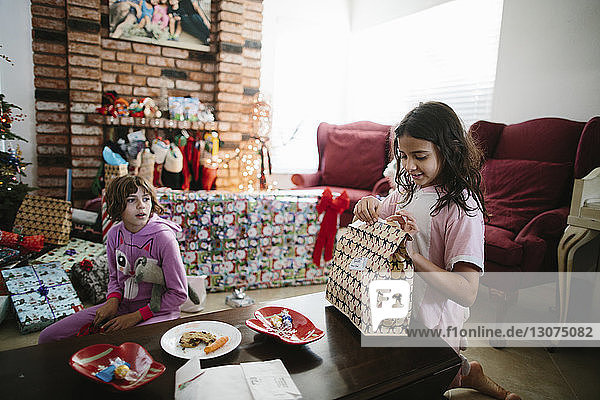 Mädchen sieht glückliche Schwester an  die ihr Weihnachtsgeschenk auf dem Tisch im Wohnzimmer öffnet