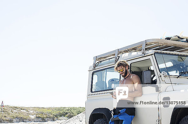 Man using mobile phone by off-road vehicle against clear sky during sunny day