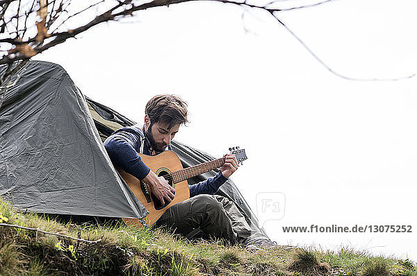 Mann spielt Gitarre vor dem Zelt auf einem Hügel vor klarem Himmel