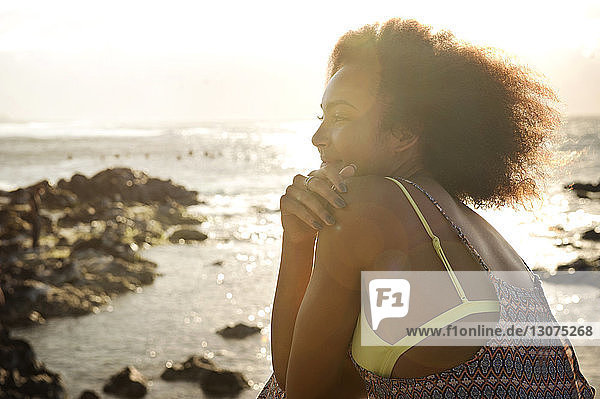 Nachdenklich lächelndes Teenager-Mädchen sitzt bei Sonnenschein am Meeresufer