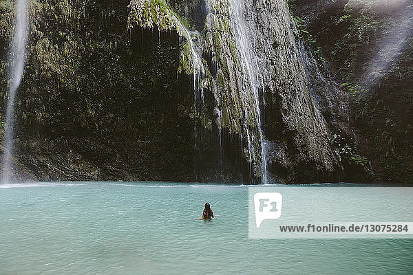 Rückansicht einer Frau  die im Fluss am Wasserfall im Wald schwimmt