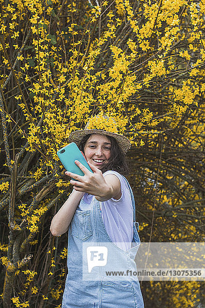 Lächelnde junge Frau  die mit einem Smartphone Selbsthilfe nimmt  während sie im Park vor gelben Blüten auf Zweigen steht
