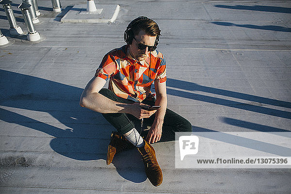 Hochwinkelansicht eines modischen Mannes  der Musik hört  während er ein Smartphone auf der Gebäudeterrasse benutzt