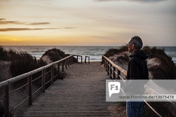 Thoughtful senior man standing on footbridge at beach during sunset