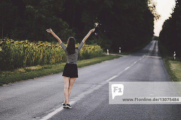 Rear view of woman with arms raised holding sparkler while standing on road