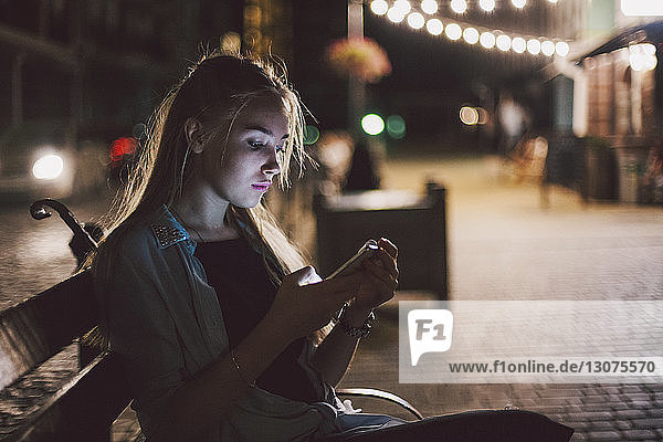 Junge Frau benutzt ein Smartphone  während sie nachts auf einer Bank am beleuchteten Bürgersteig in der Stadt sitzt