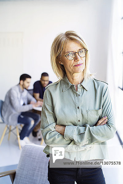 Thoughtful businesswoman looking away while standing in office with colleagues in background