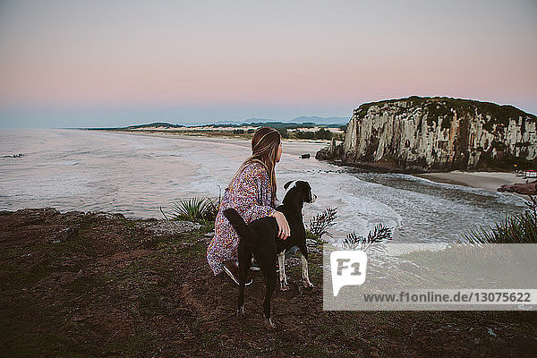 Side view of woman crouching by dog on hill at beach during sunset