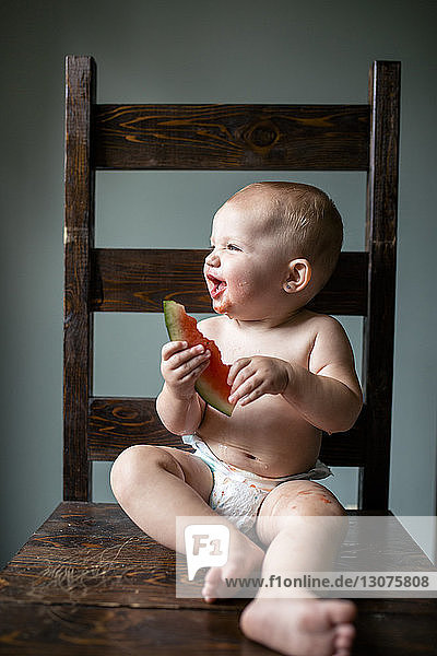 Fröhliches kleines Mädchen schaut weg  während es zu Hause einen Wassermelonenscheibenstuhl an die Wand hält