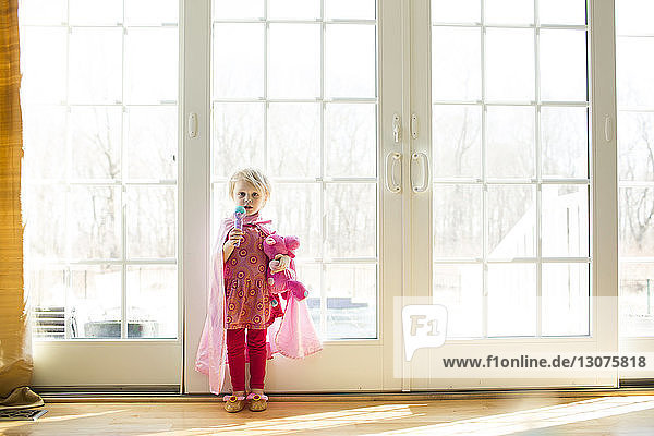 Portrait of girl wearing cape holding toys standing by window at home