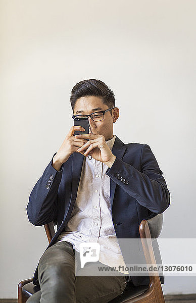 Geschäftsmann benutzt Mobiltelefon  während er im Büro auf einem Stuhl an der Wand sitzt