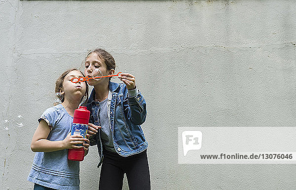 Sisters blowing bubbles while standing against wall
