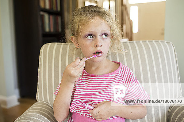 Mädchen trägt Lipgloss auf  während sie zu Hause auf einem Sessel sitzt