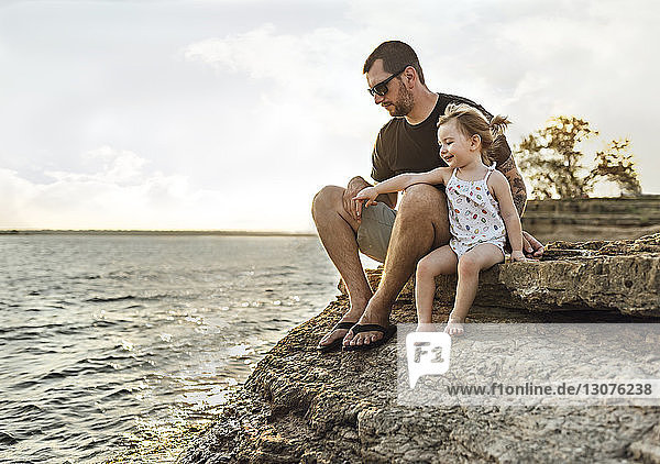 Fröhliche Tochter zeigt auf Fluss  während sie neben dem Vater auf Felsen sitzt