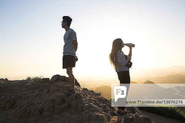 Woman drinking water while man looking at view while standing on mountain