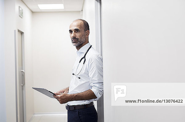 Doctor with tablet computer looking away while standing by door in corridor