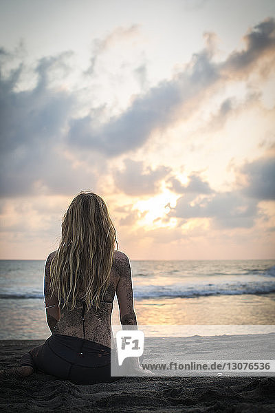 Rear view of woman sitting on shore at beach against sky during sunset