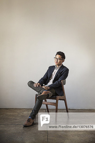 Lächelnder selbstbewusster Geschäftsmann in voller Länge auf Stuhl sitzend an Wand im Büro