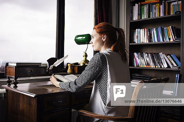 Seitenansicht einer nachdenklichen Geschäftsfrau  die ein Buch in der Hand hält und am Schreibtisch im Kreativbüro sitzt