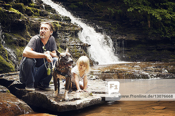 Grossvater und Enkelin mit Hund kauern auf Felsen am Seeufer im Wald