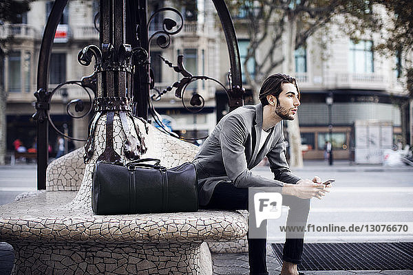 Man with mobile phone looking away while sitting on bench