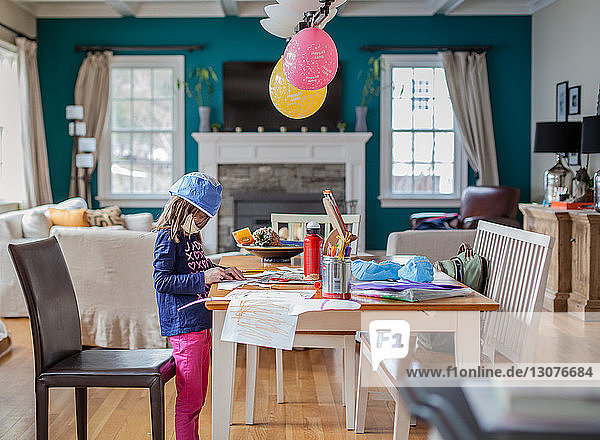 Seitenansicht eines Mädchens mit Schutzmaske und Kappe beim Zeichnen am Tisch zu Hause