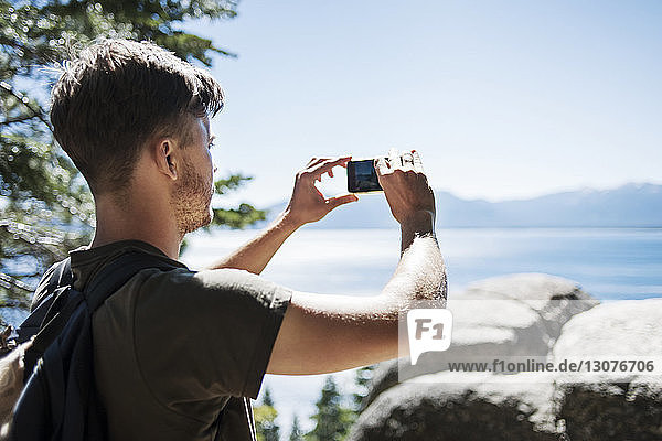 Rear view of man photographing sea through smart phone against clear blue sky